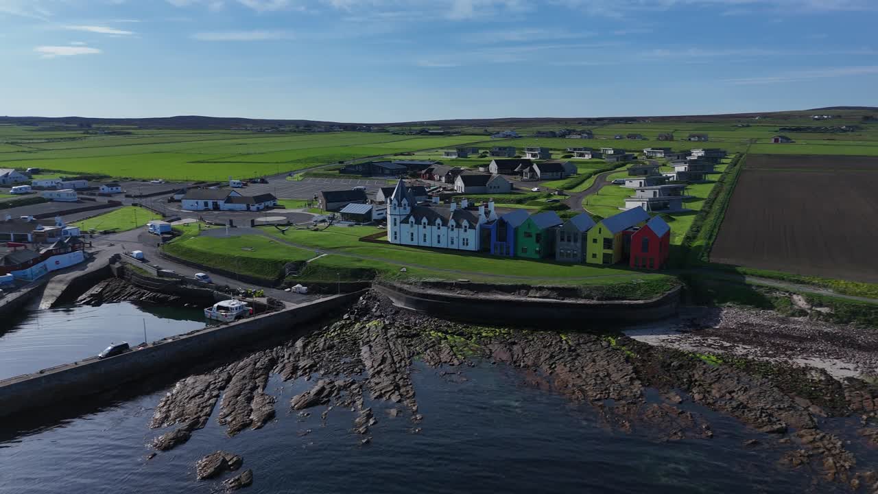 Panning shot of John O'Groats harbour with colourful buildings next to the signpost