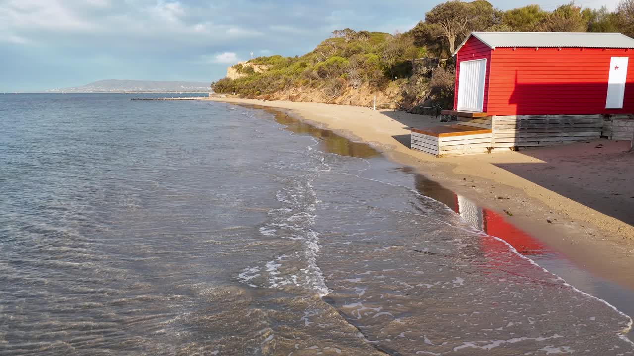 Calm sea waves approach sandy beach beside vibrant red bathing box under soft natural daylight