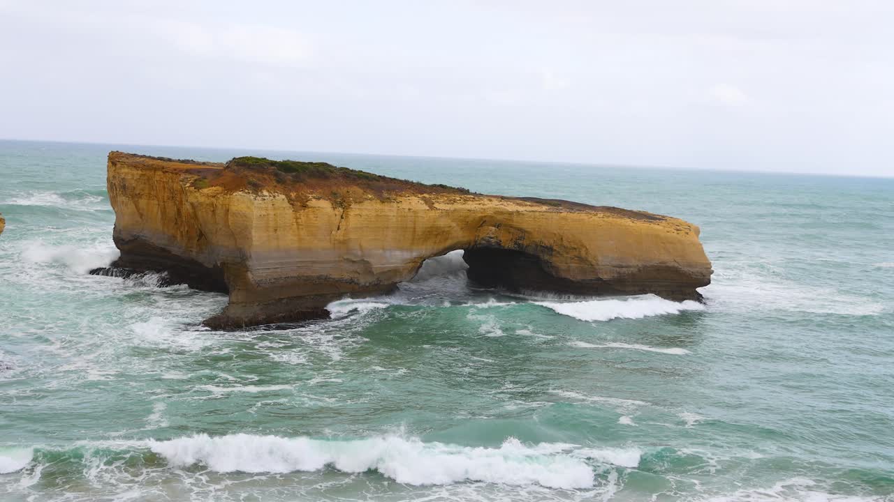 Dynamic ocean waves crash against the London Bridge rock formation under overcast skies at Port Campbell, Australia