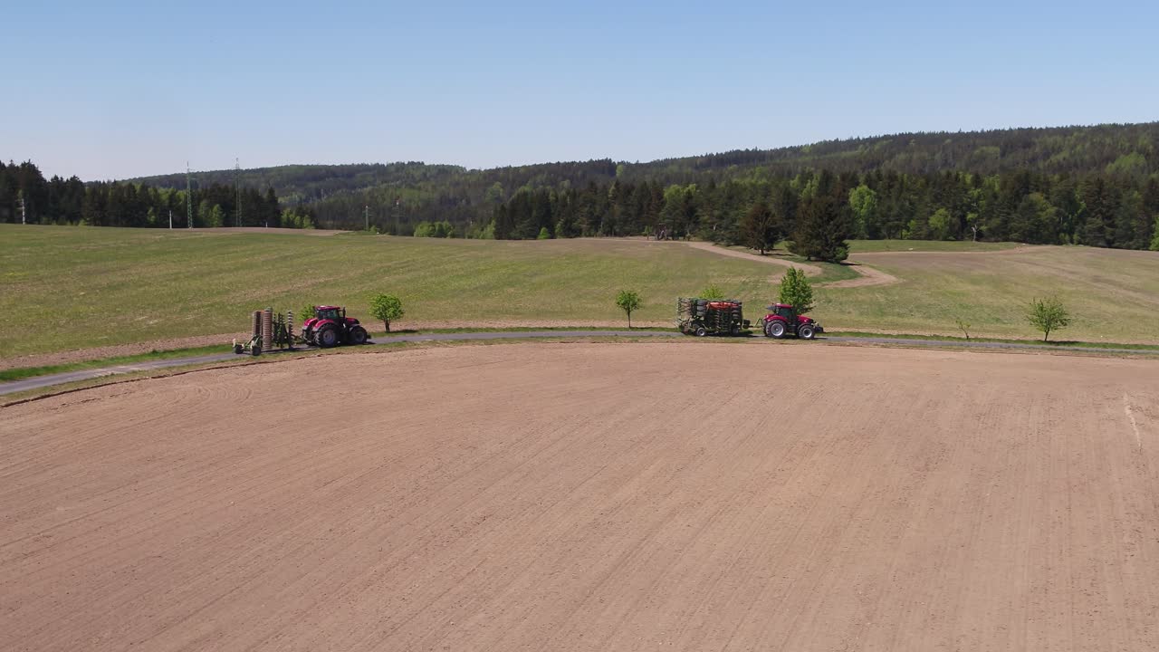 Drone pan right showing red tractors moving through winding road surrounded by farmland and trees