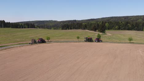 drone pan right showing red tractors moving through winding road surrounded by farmland and trees