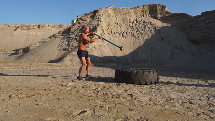 Muscle athlete strongman man hits a hammer on a huge wheel in the sandy mountains in slow motion. strength and endurance training for wrestlers