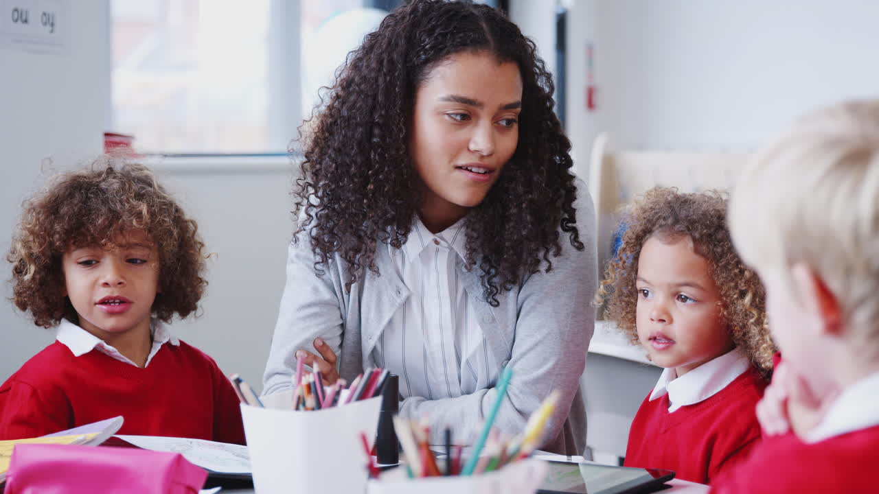 Smiling female infant school teacher sitting at a table in class with schoolchildren, waist up