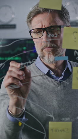 Mature-investment-banker-draws-on-glass-wall-with-sticky-notes-in-broker-agency-office.-Businessman-develops-and-analyzes-company-business-strategy.-Colleagues-work-on-computers-with-real-time-stocks.