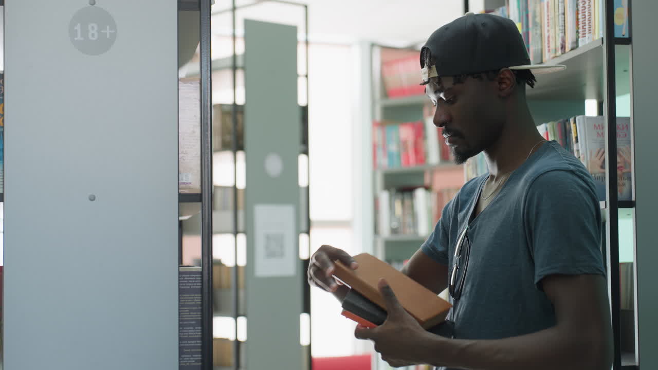 Side view of college student in cap retrieving book from shelf in bright modern library, holding black hardcover, sunglasses hanging on shirt, focused expression, rows of books