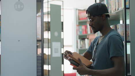 Side view of college student in cap retrieving book from shelf in bright modern library, holding black hardcover, sunglasses hanging on shirt, focused expression, rows of books