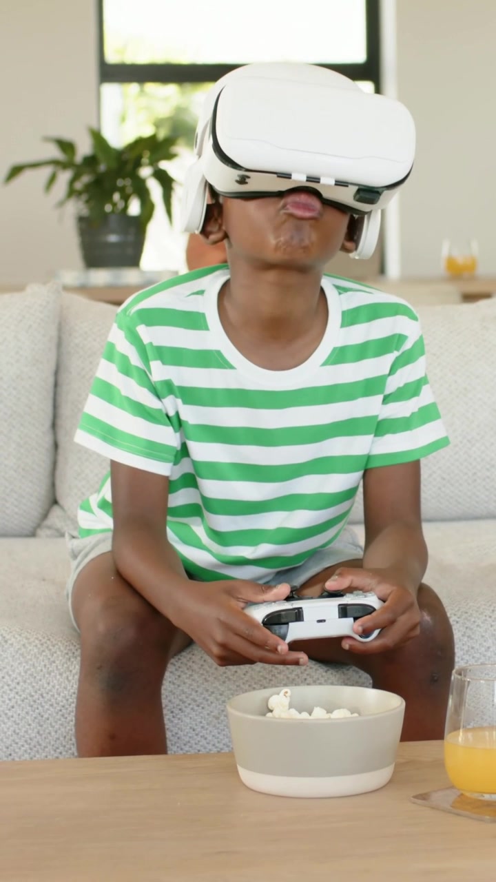 Vertical video: Game starting, child in striped shirt wearing VR headset using pad on couch at home