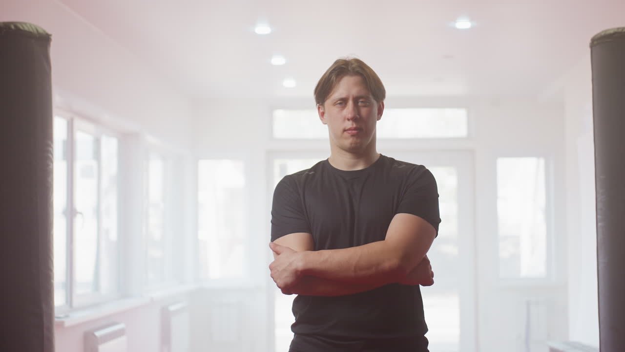 Two fighters in black sportswear stand on red training mat inside gym, preparing for combat practice, discussing strategy with serious focused expressions surrounded by punching bags and training equipment