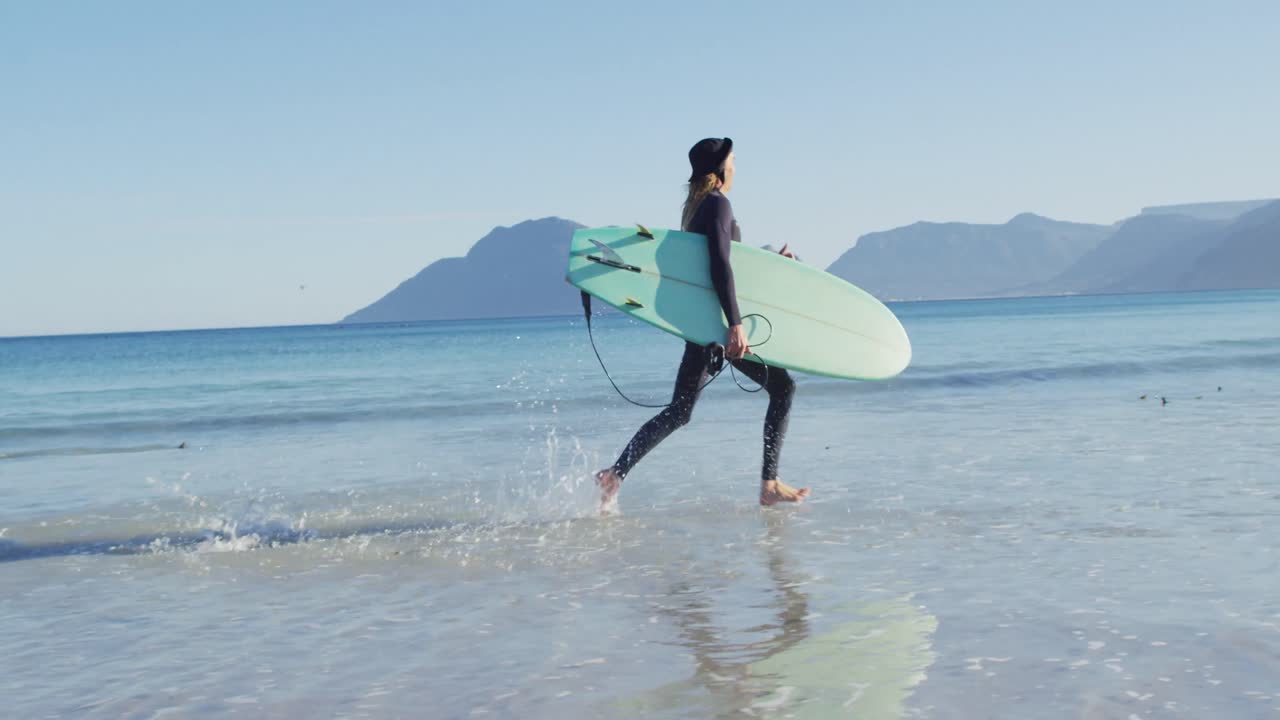video de un hombre caucásico con rastas en traje de neopreno llevando una tabla de surf corriendo en el mar en una playa soleada