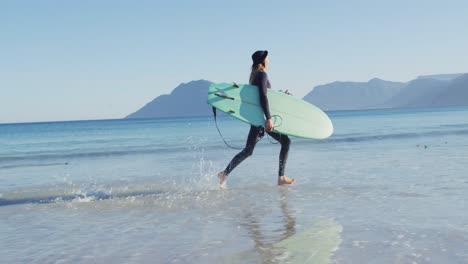 Video-Eines-Kaukasischen-Mannes-Mit-Dreadlocks-Im-Neoprenanzug,-Der-Ein-Surfbrett-Trägt-Und-Am-Sonnigen-Strand-Im-Meer-Läuft