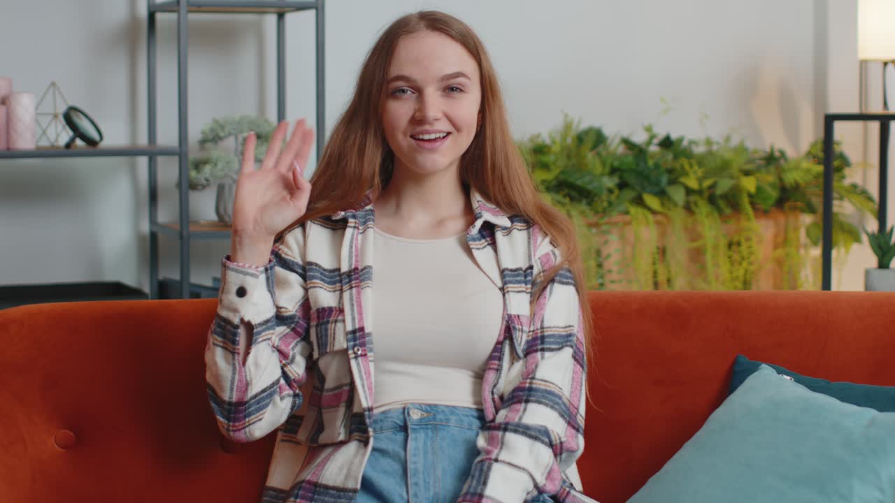 Young woman smiling friendly at camera waving hands gesturing hello hi greeting at home on sofa