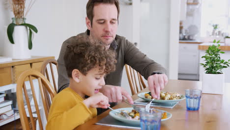 Alleinerziehender-Vater-Hilft-Seinem-Sohn-Beim-Schneiden-Von-Essen,-Während-Sie-Zu-Hause-Am-Tisch-Sitzen-Und-Essen