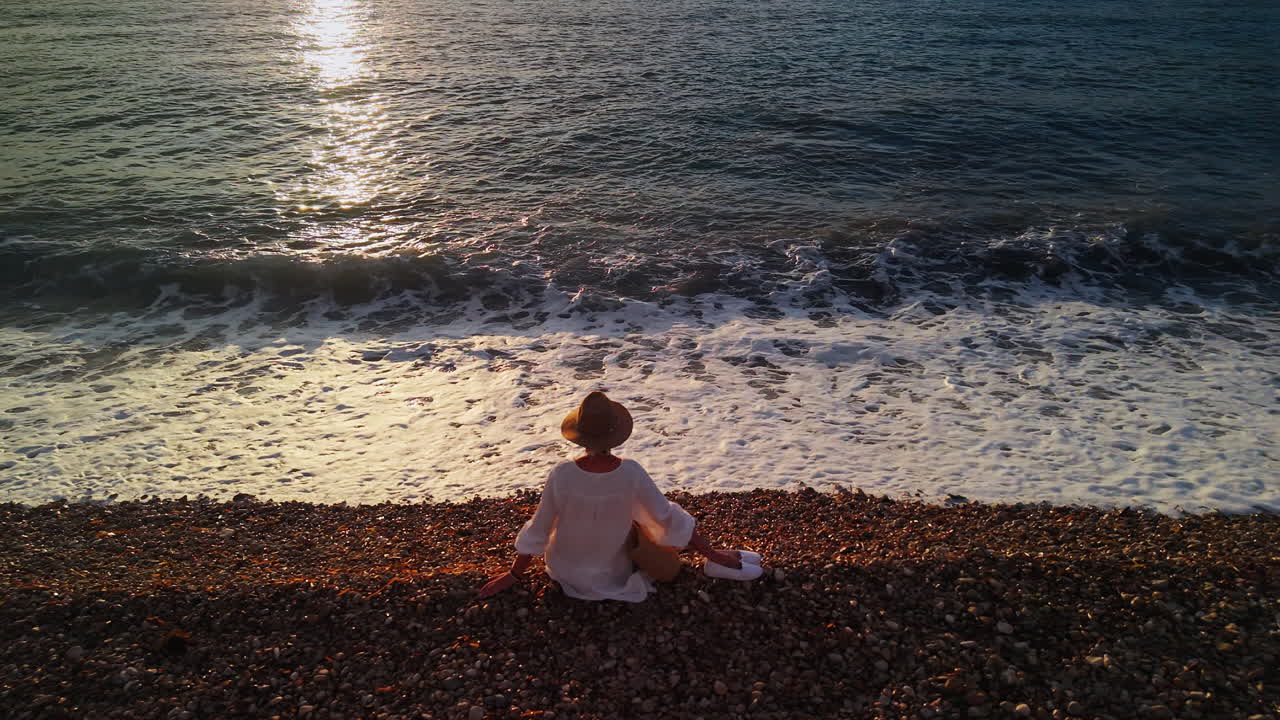 Donna seduta su una spiaggia di ciottoli che osserva le onde al tramonto