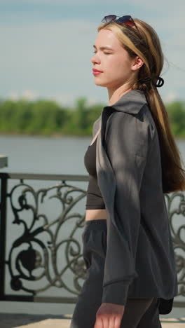 lady walking near riverfront with shopping bag in hand, passing a decorative metal fence, background features blurred bridge, greenery, and clear sky on a sunny day, while she gazes towards the river