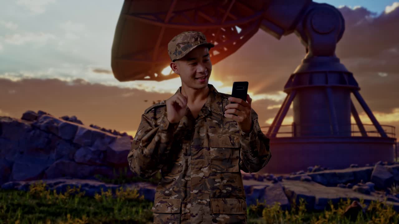 Soldato asiatico che saluta con la mano durante una videochiamata sullo smartphone mentre è in piedi accanto a un'antenna parabolica.