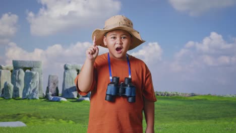 asian boy with a hat and binoculars thinking and looking around then raising his index finger while traveling in stonehenge. boy researcher, travel tourism adventure concept