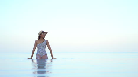 A-young,-fit-woman-in-a-bathing-suit,-sunglasses,-and-sun-hat-sits-on-the-invisible-edge-of-a-resort-swimming-pool