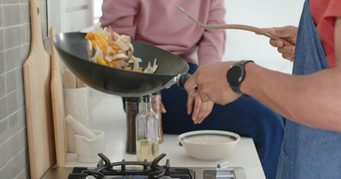 Fiamma a gas che riscalda un wok nero, cuoco che salta le verdure con una spatola sul fornello a gas.