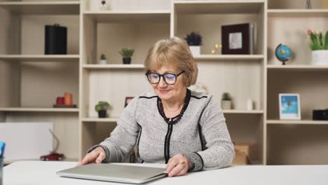 older woman removes her glasses and finishes work on the laptop, smiling as she looks into the camera