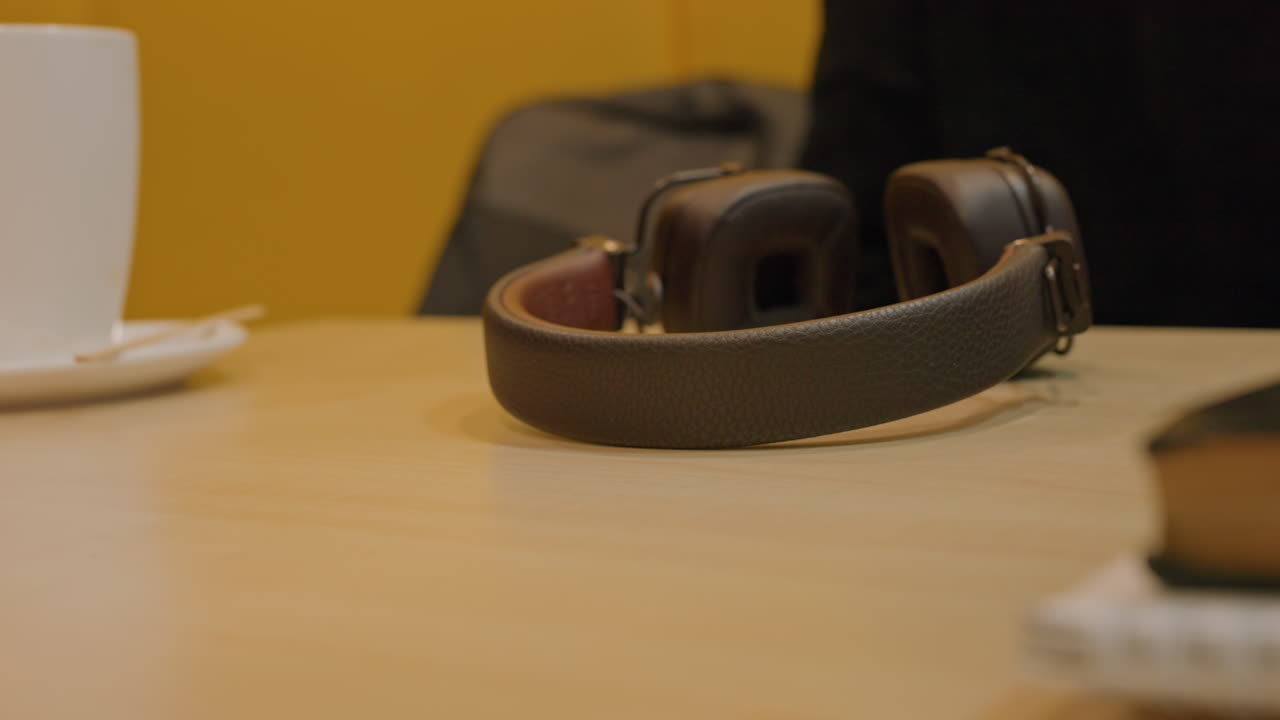 Tangan meraih headphone dengan orang berkulit hitam di meja, meletakkan headphone kulit di permukaan kayu, cangkir kopi dan buku di tepi, latar belakang kuning hangat, gestur taktil, ritual pra-rekaman yang tenang