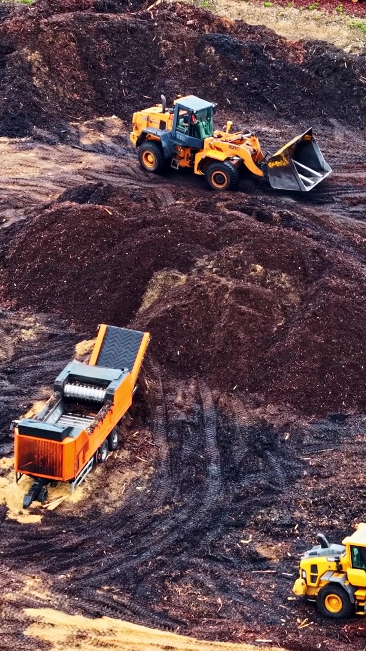 Vertical aerial parallax over yellow wheel loader and orange wood chipper processing bark mulch into sawdust beside stacked logs at timber sawmill yard in Latvia