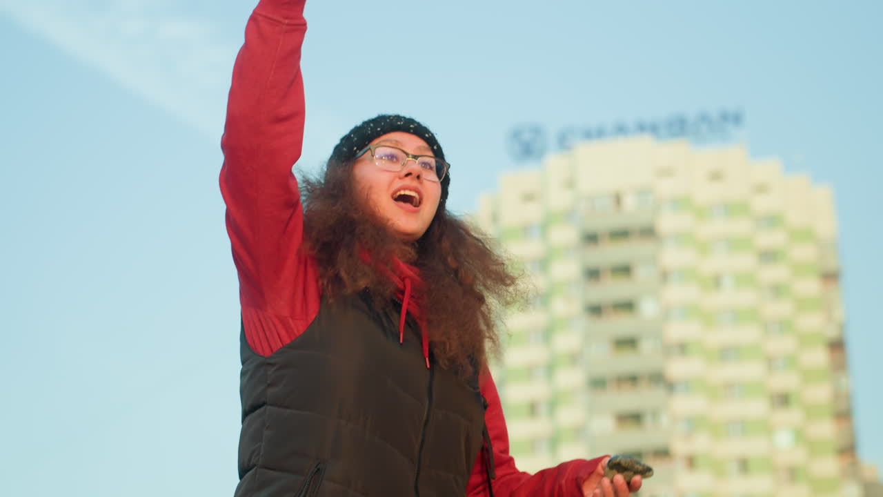 Donna all'aperto vicino a una torre che controlla un drone durante l'ora d'oro, che indossa berretto e gilet, che tiene il telecomando mentre sorride e gesticola, cielo azzurro limpido e edificio residenziale sullo sfondo, rilassata