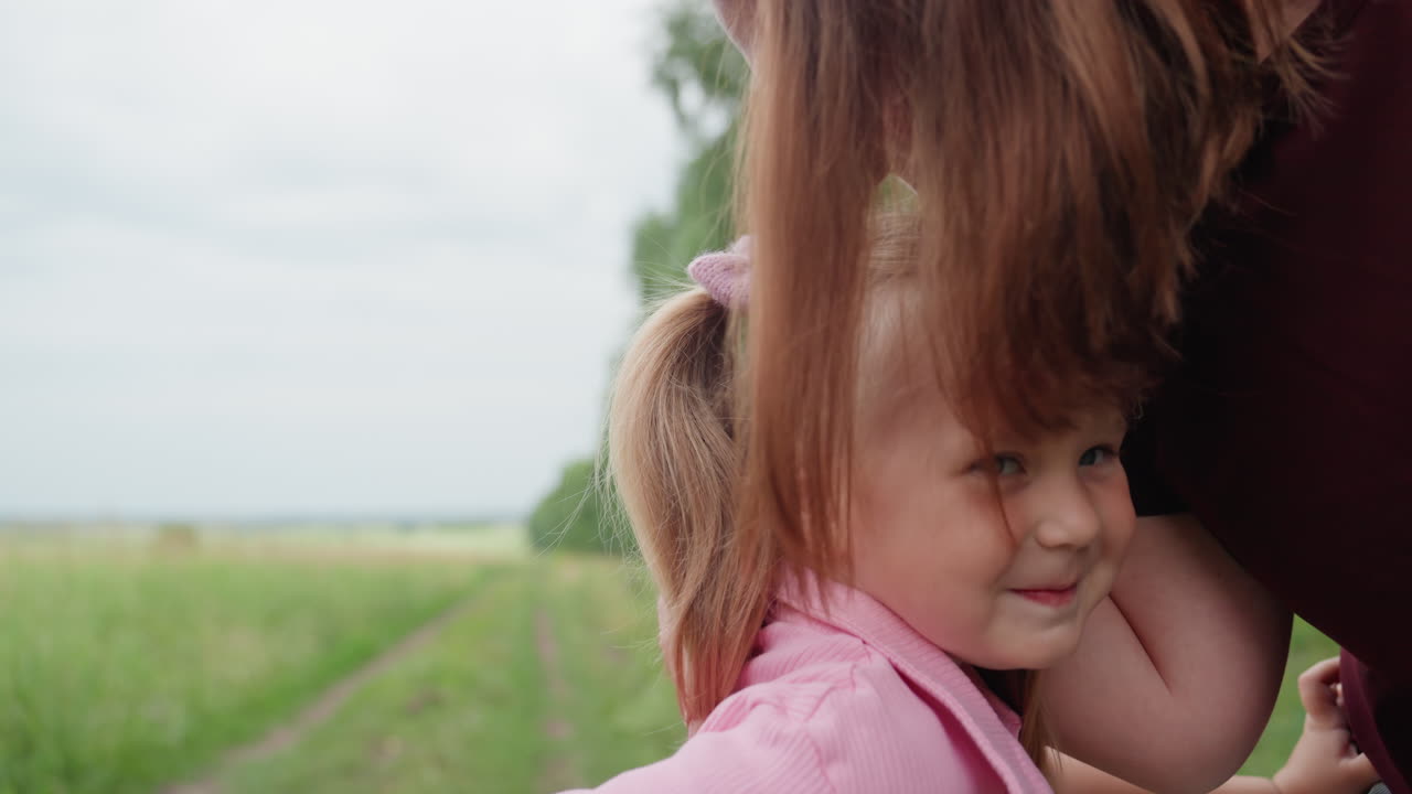 Niña riendo en un campo de hierba, polo rosa, coleta, hermanos cogidos de la mano, madre acomodándose el pelo, cálido día de campo familiar, momento espontáneo y afectuoso, risas y tirón juguetón en una pradera verde.