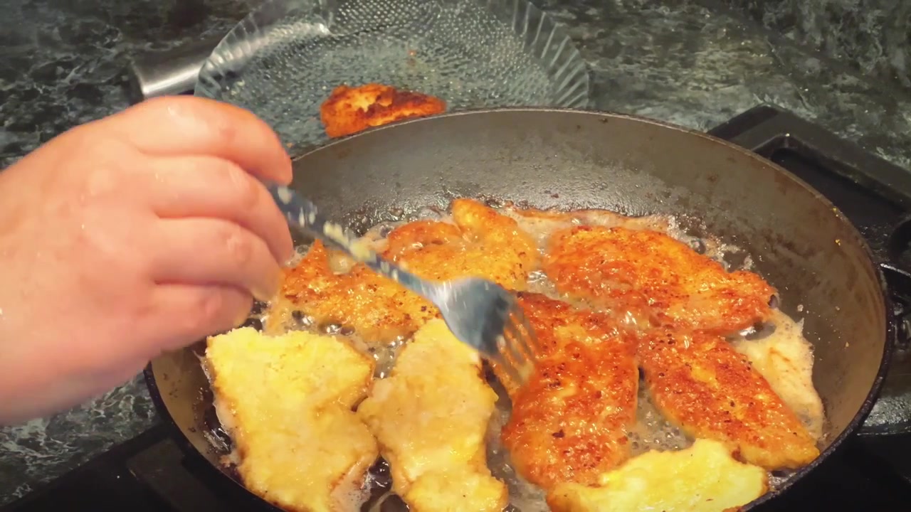 Primo piano di una mano che usa una forchetta per girare petti di pollo impanati e conditi che friggono in olio caldo all'interno di una padella. Ideale per tutorial di cucina e promozioni di ricette.