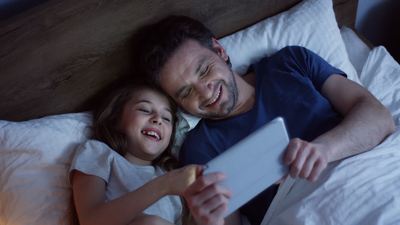 Top view of little girl lying on the bed with her father and watching video on the tablet, laughing and having fun