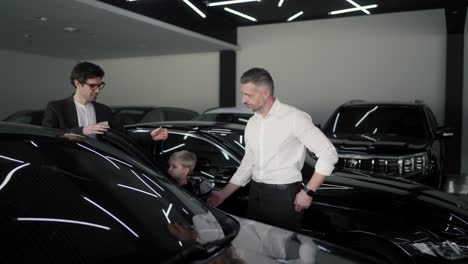 A-confident-brunette-guy-in-a-business-suit-an-assistant-at-a-car-dealership-communicates-with-a-middle-aged-man-and-lets-his-little-son-get-into-the-car