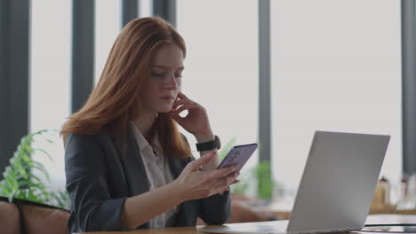 Junge-Geschäftsfrau-Nutzt-Smartphone-Mit-Laptop.-Geschäftsfrau-In-Einem-Modernen-Büro-Mit-Panoramafenster,-Sonniger-Tag,-Laptop,-Tablet,-Nutzung-Eines-Mobiltelefons-In-Einer-Situation