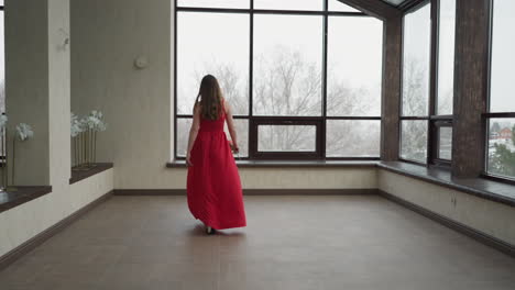 model gracefully strolls along studio. young woman dressed in stylish wine-colored gown holds rose in hand in gallery with panoramic windows