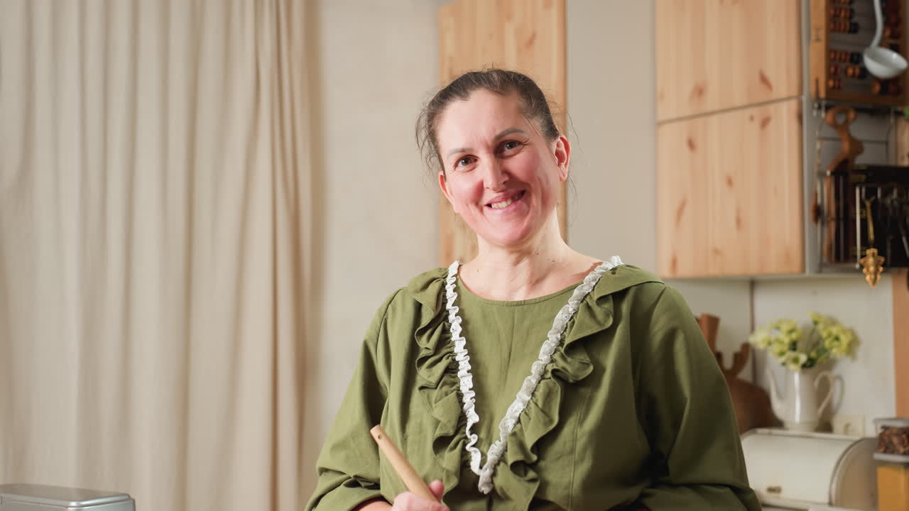 Cheerful cook in green gown with white lace trim standing in kitchen, holding spatula with head playfully tilted and bright smile, surrounded by cozy decor, wooden cabinets, and kitchen appliances