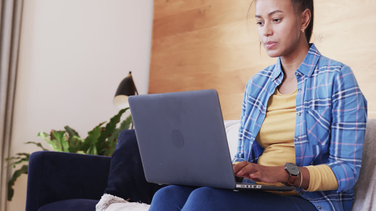 Focused biracial woman sitting on couch using laptop in living room, in slow motion