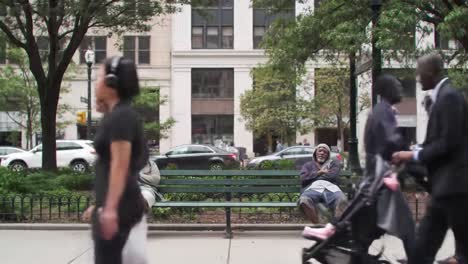 an elderly man sits comfortably on a bench in the park, observing the bustling crowd around him. people walk by, some with strollers, others engaged in conversations, creating a lively atmosphere.