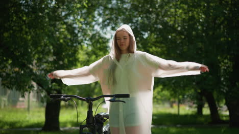 young woman in translucent raincoat stretches her arms joyfully near bicycle, swaying her hands in sunny park surrounded by lush greenery