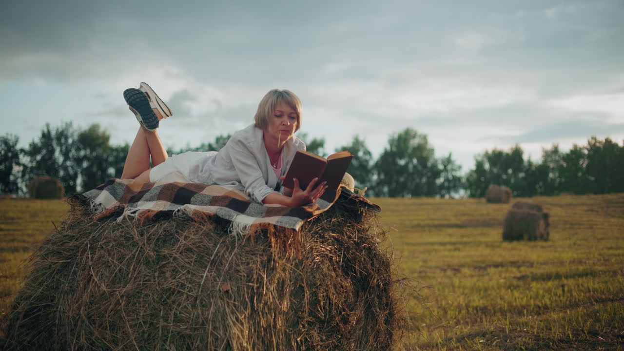 Женщина с ногой, поднятой на стог сена с клетчатым пледом, читает книгу на открытом поле с соломинкой во рту, наслаждаясь мирным моментом на природе в окружении природы.