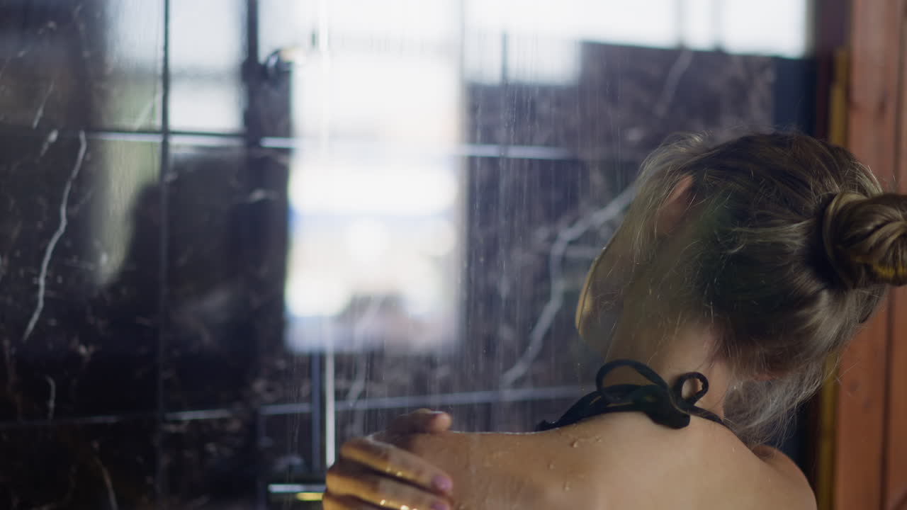 젊은 여성이 샤워 객실에서 뜨거운 물  ⁇ 에서 어깨를  ⁇ 고 있습니다.