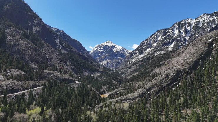 Drone che vola lentamente attraverso montagne rocciose e frastagliate con piccole chiazze di neve, alta cima montuosa sullo sfondo coperta di neve, Trico Peak, Monti San Juan, Ouray Colorado