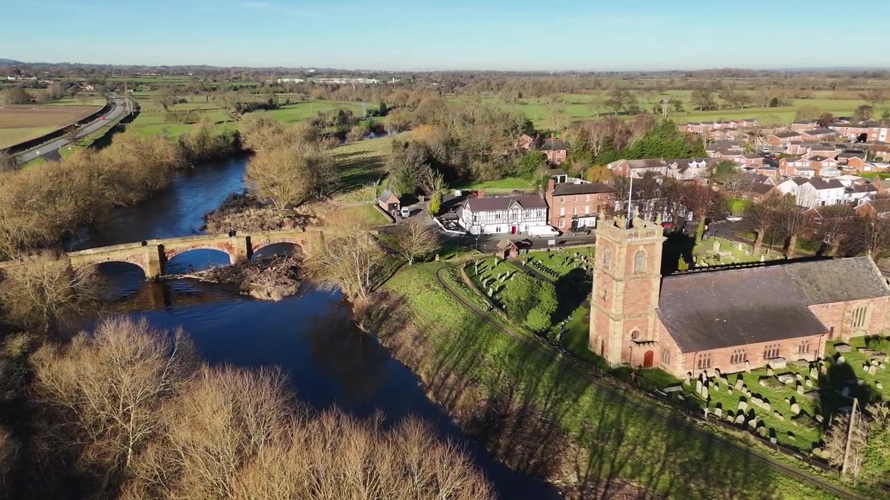 Vista aérea de drone de uma ponte de pedra histórica cruzando um rio tranquilo ao lado de uma igreja paroquial tradicional na zona rural inglesa. Cercada por campos verdes, árvores e paisagem rural, esta cena