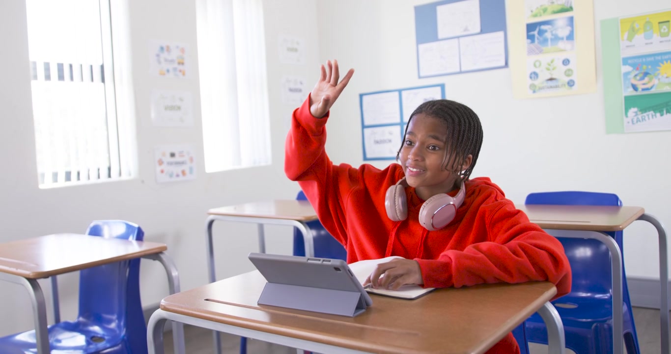 Adolescente afro-americano fazendo anotações na carteira da sala de aula, verificando o tablet e levantando a mão para pedir ajuda