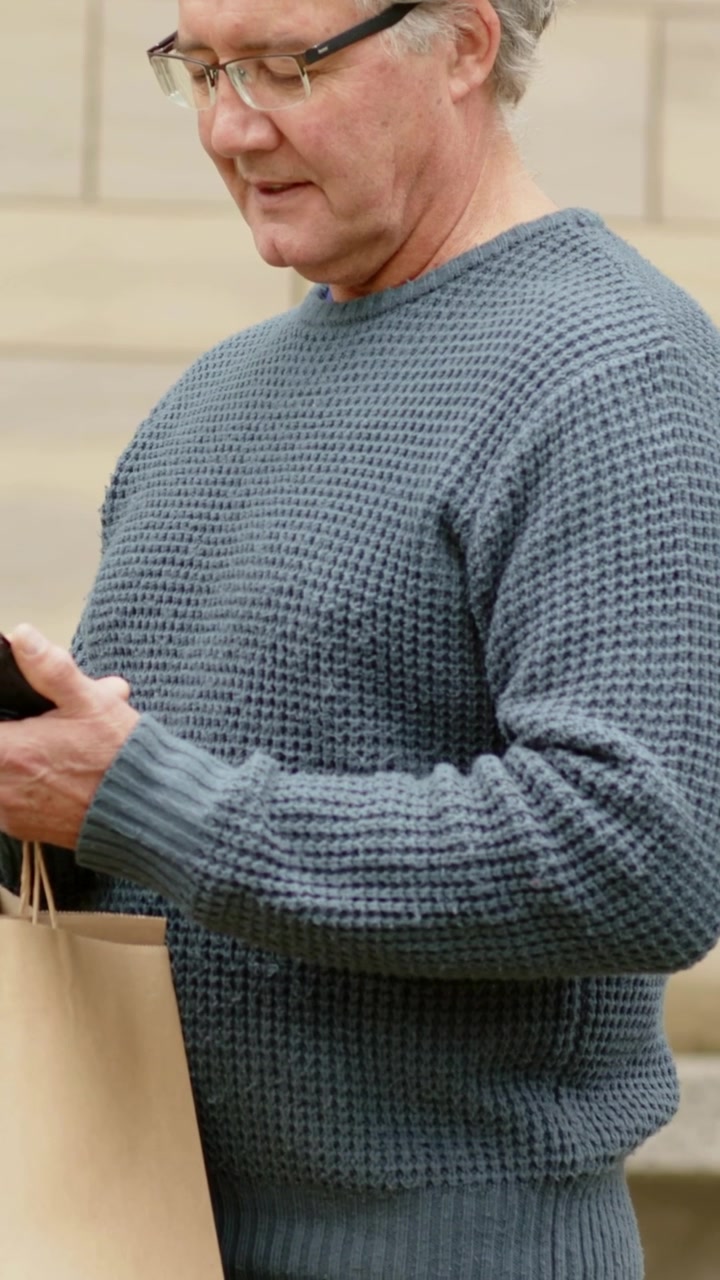 Video verticale: Anziano con gli occhiali che guarda attentamente il telefono e digita, alzando lo sguardo per scansionare i gradini mentre tiene una borsa.
