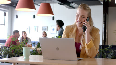 Happy-caucasian-casual-businesswoman-talking-on-smartphone-using-laptop-in-office,-slow-motion