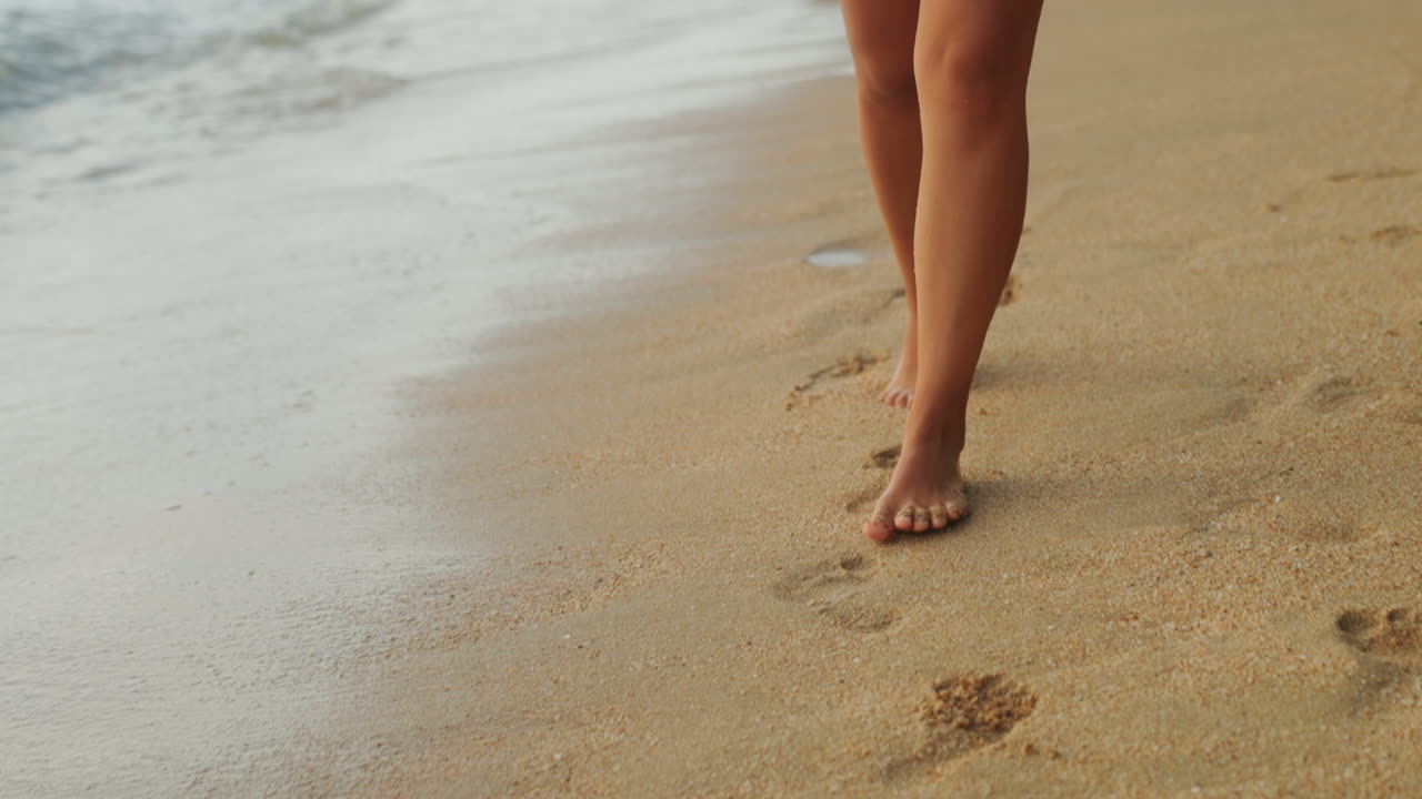Passeggiata tranquilla in spiaggia. Donna che cammina silenziosamente sulla sabbia della spiaggia all'alba. Calma passeggiata solitaria lungo il litorale sabbioso illuminato dal sole al mattino presto. Pacifica passeggiata mattutina in una zona costiera sabbiosa e deserta baciata dal sole.
