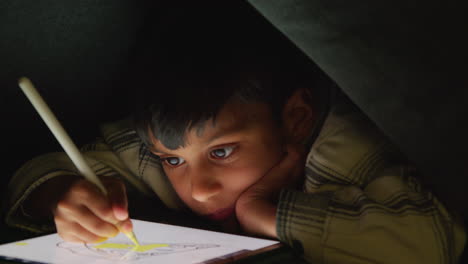 close up of young boy in home made camp made from cushions playing and drawing with digital tablet and stylus at night