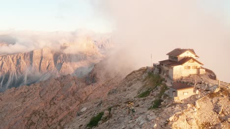 Epic-Drone-shot-featuring-mountain-hut-on-the-peak-covered-by-clouds-during-sunset---Rifugio-Nuvolau-at-Cinque-Torri,-Dolomites