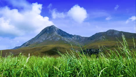 Zeitraffer---Wolken-Ziehen-über-Berggipfel,-Grünes-Feld-Im-Vordergrund,-Atemberaubende-Landschaft