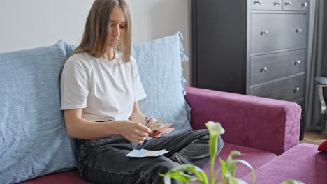 woman sitting on couch while counting euro cash bills from her hand towards her knees at home, static