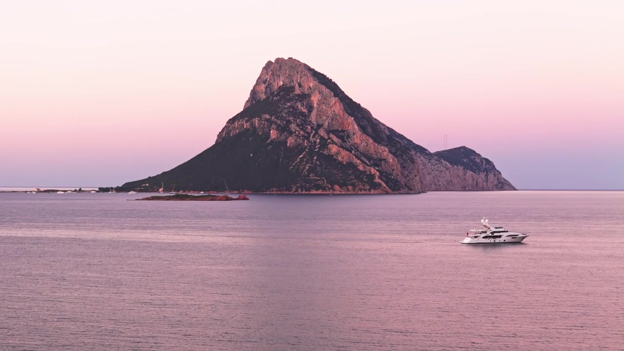 Majestætiske ø Tavolara på Sardinien, Italien, tårner sig op over det rolige Middelhav. En luksusyacht sejler under en magisk lyserød solnedgang