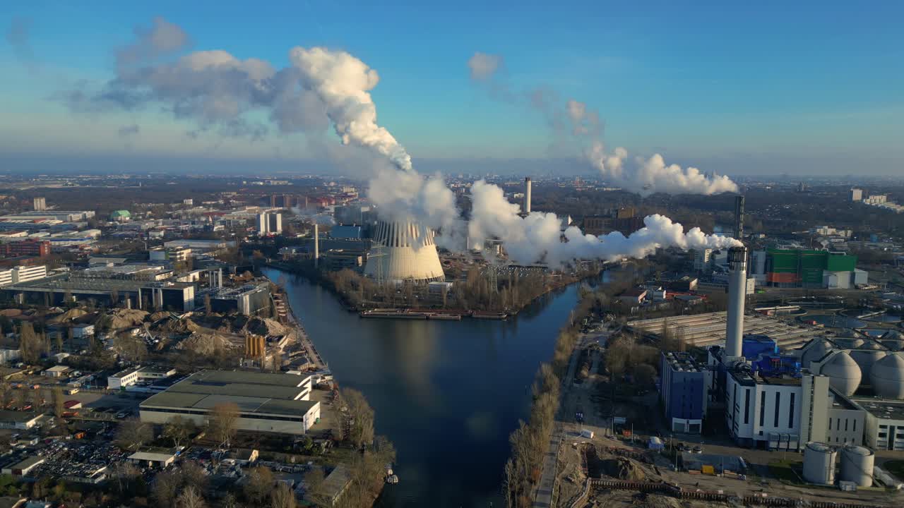 Aerial view of a thermal power plant emitting smoke on a sunny day, with a river and a city in the background. Smooth aerial view flight panorama overview drone